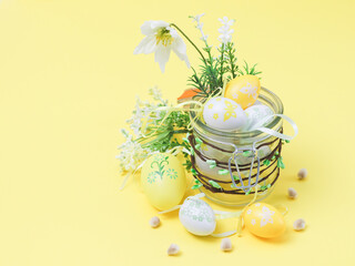 A glass jar with buds in which are decorative Easter eggs, spring flowers and willow buds on a yellow.
