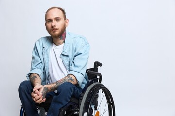 A man in a wheelchair looks thoughtfully at the camera, copy space, with tattoos on his arms sits...