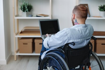 A man in a wheelchair freelancer works at a laptop at home view from the back, copy space, integration into society, health concept man with disabilities, real person