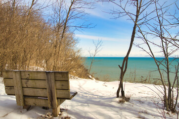 Beneath a mostly clear Winter sky, an empty wooden bench faces the expanse of Lake Michigan from atop a snowy forested bluff at Lion's Den Gorge near Grafton, WI.