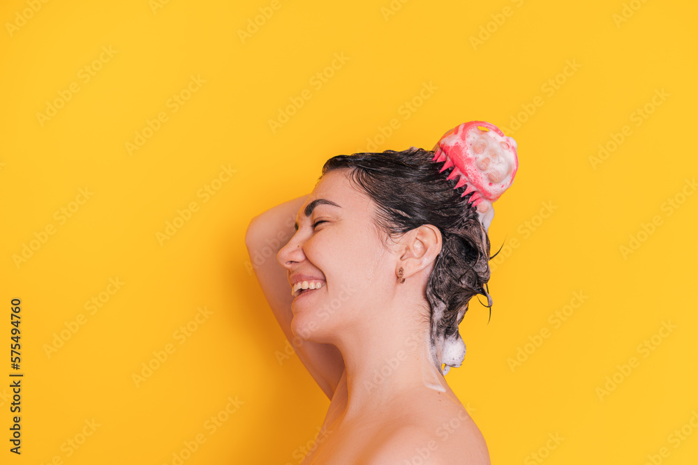 Wall mural Lady with soapy head takes care of the scalp with massager in pink against yellow background.