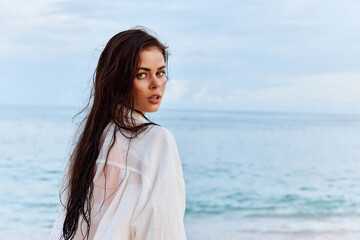 Portrait of a beautiful pensive woman with tanned skin in a white beach shirt with wet hair after swimming on the beach ocean sunset light with clouds