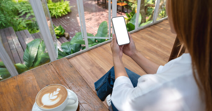 Mockup image of a woman holding and using mobile phone with blank desktop screen with coffee cup on the table in cafe