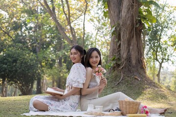 Charming beautiful young Asian woman in a lovely dress, holding a flower bouquet, enjoying a picnic with her friend in the park