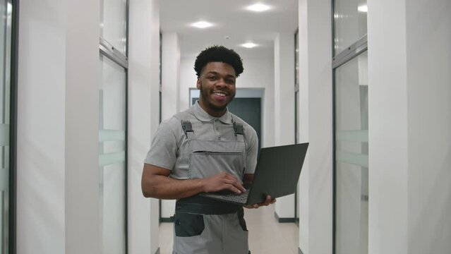 African CCTV cameras installer in uniform stands in corridor, smiles and looks at camera. He sets up security cameras in office using software on laptop. Concept of surveillance system. Dolly zoom.