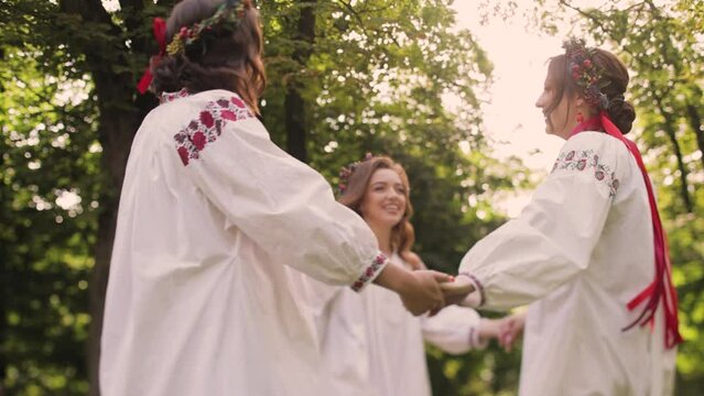 Three women in round dance roundelay. Girls wear Ancient ethno Ukrainian embroidered shirt dress. Bride in a wreath of flowers. Medium shot