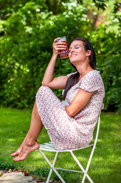 A 38 year old brunette woman outdoors sitting on a chair wearing a summer dress and holding a glass of iced tea.