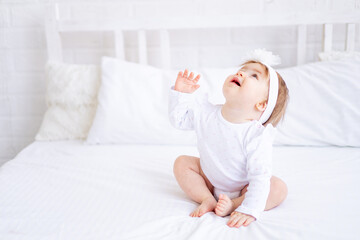 funny happy baby girl sitting on the bed in white clothes and raising her hands looking up, a small child on a cotton bed at home smiling, the concept of children's goods