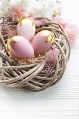 Naklejka na ściany i meble Easter eggs painted pink and gold and feathers in a nest on a white wooden background.