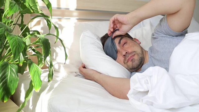 Sleepy man with a sleep mask in bed in his room. Didn't get enough sleep and continues to sleep.