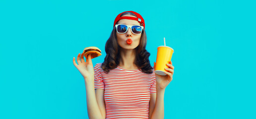 Portrait of stylish young woman drinking juice with burger fast food on blue background