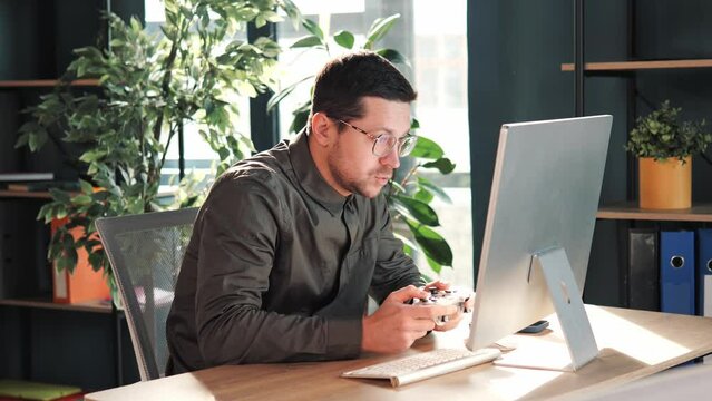 Funny confused office worker happy man employee gamer hold controller joystick playing video game on computer. The man looks around to see if anyone is there. Male gamer having fun free time at work.