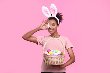 Happy African American woman in bunny ears headband covering eye with Easter egg on pink background