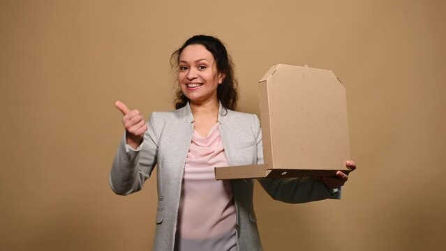 Beautiful happy woman opens a cardboard flat box with hot Italian pizza, shows thumb up and tasty gesture, kisses fingers and says bellissimo, isolated on beige background. Gourmet culinary concept