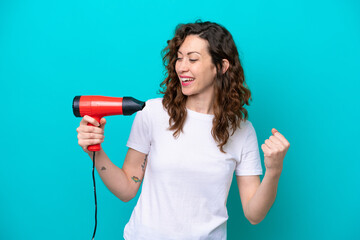 Young caucasian woman holding a hairdryer isolated on blue background celebrating a victory