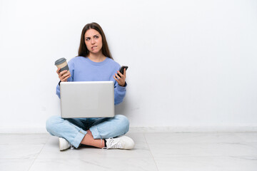Young caucasian woman with laptop sitting on the floor isolated on white background holding coffee to take away and a mobile while thinking something