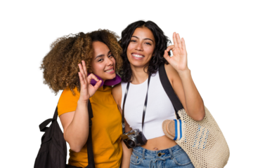 Two diverse friends on a beach vacation with vintage camera, beach bag, and travel pillow cheerful and confident showing ok gesture.