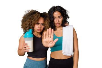 Two best friends are ready to go to the gym, one of them holds a protein shake, standing with outstretched hand showing stop sign, preventing you.