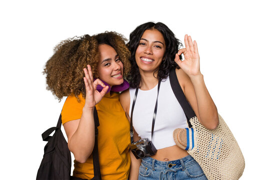 Two diverse friends on a beach vacation with vintage camera, beach bag, and travel pillow cheerful and confident showing ok gesture.