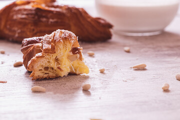 A bitten croissant on a wooden table with a whole croissant and a bowl of milk in the background