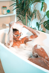 Obraz na płótnie Canvas Excited woman with sunglasses playing with water and foam in bathtub. Happiness and wellbeing concept. High quality photo