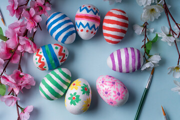 Vibrant and cheerful flat lay background features circle of colorful Easter eggs adorned with festive decorations, surrounded by spring blossoms and brushes. For website, social media, greeting cards