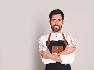 Smiling hairdresser wearing apron holding scissors on light grey background, space for text