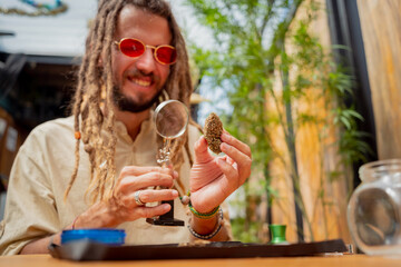 Hippie style man examines under a magnifying glass the joints and buds of medical marijuana