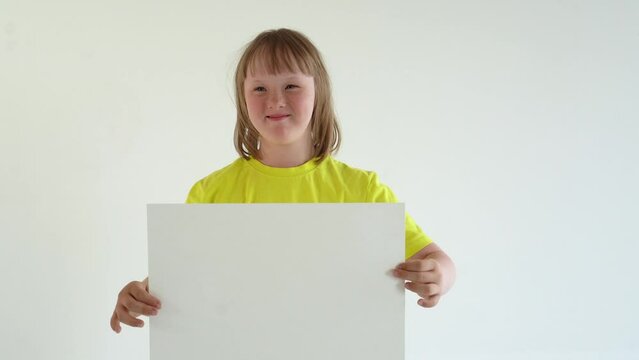 A teenage girl with blond hair in a yellow T-shirt holds a sheet of A3 paper and smiles. mock up and space for text, Down syndrome awareness concept. International Down Syndrome Day.