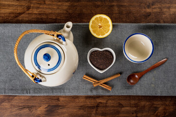 Porcelain teapot next to a small heart-shaped bowl with ground tea, a cup, half a lemon and pieces of cinnamon, top view.