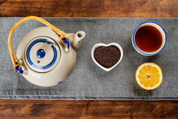 Porcelain teapot next to a small heart-shaped bowl with ground tea, a cup with tea and half a lemon, top view.