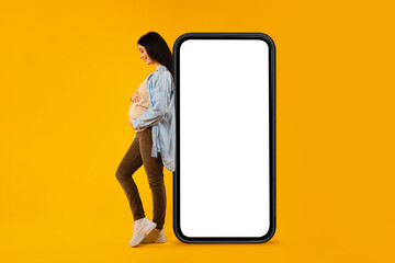 Happy pregnant woman leaning on huge blank cellphone and hugging her belly, advertising mobile app...
