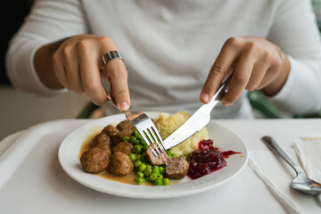 Close up on hands of unknown caucasian man sit at the table with fork and knife eat meatballs with peas and mashed potato
