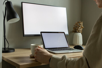 Person working  from home on laptop with larger monitor in the background and holding a coffee mug.