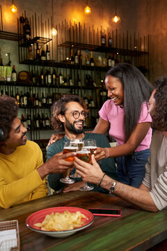Vertical photo of group co-workers toast with beer at the restaurant bar after work at the pub happy hour. Happy friends having fun together drinking alcohol indoors. People enjoying their free time.