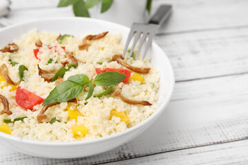 Bowl of delicious couscous with vegetables and basil served on white wooden table, closeup. Space for text