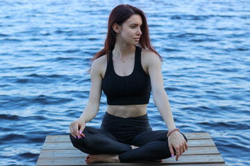 Fototapeta na wymiar A beautiful attractive girl practices yoga in the lotus position near the lake. Relaxing photo in warm colors.