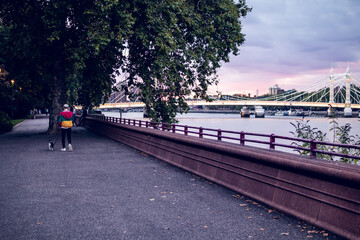 Naklejka premium Chico joven paseando un perro en un paisaje urbano de un parque al lado del río Támesis de Londres.