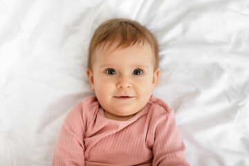 Adorable caucasian baby in bodysuit lying on bed at home, resting on white bedsheets, closeup portrait, top view