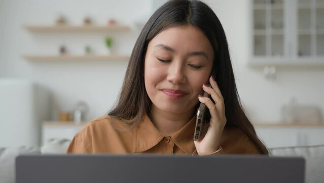 Female Asian entrepreneur korean businesswoman woman freelancer sit on couch negotiate by mobile phone speaking cellphone working multitask with laptop search data e-learning answer call business talk