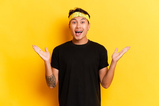 Positive funny young ethnic man with headband and pierced ear spread palms aside has amazed expression smiles with wide opened mouth wears black casual t-shirt isolated next to yellow background