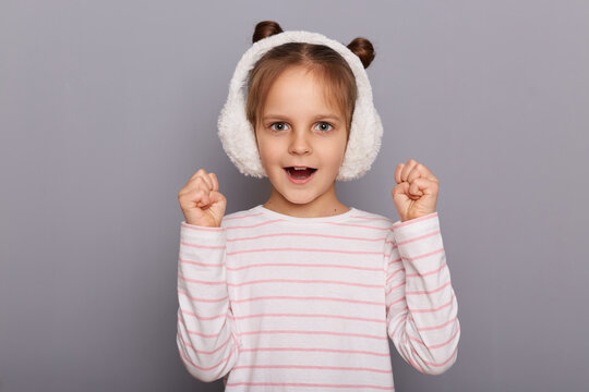 Portrait of extremely happy surprised cute little girl wearing fur earmuffs and striped shirt standing isolated over gray background, standing with clenched fists, screaming with happiness.