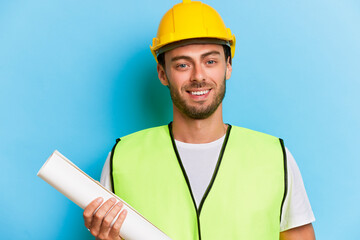 People and work concept. Photo of positive construction male worker wears hardhat and uniform holds blueprint smiles gladfully to the camera isolated over blue background