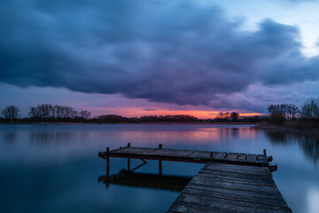 Obraz premium View from a wooden pier on a calm lake during sunset with dark clouds