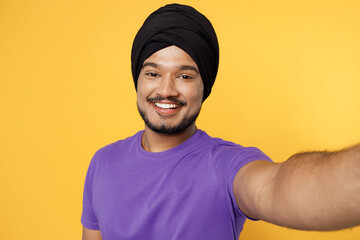 Close up smiling happy fun devotee Sikh Indian man ties his traditional turban dastar wear purple t-shirt do selfie shot pov on mobile cell phone isolated on plain yellow background studio portrait