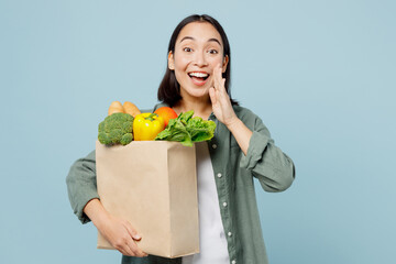 Fototapeta na wymiar Young woman in casual clothes hold brown paper bag with food products scream share hot news hands near mouth isolated on plain blue background studio portrait Delivery service from shop or restaurant