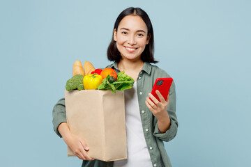 Fototapeta na wymiar Young smiling woman wear casual clothes hold brown paper bag with food products use mobile cell phone isolated on plain blue cyan background studio portrait Delivery service from shop or restaurant.