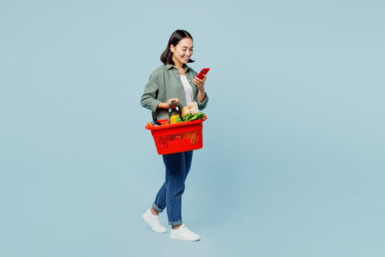 Full body side view fun young woman wear casual clothes hold red basket with food products use mobile phone isolated on plain blue background studio portrait. Delivery service from shop or restaurant.