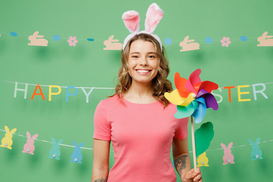 Young smiling happy cheerful woman wear pink casual clothes bunny rabbit ears hold in hand toy windmill isolated on plain pastel light green background studio portrait. Lifestyle Happy Easter concept.