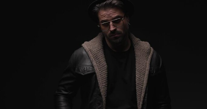 confident bearded man with hat and glasses looking to side and adjusting leather jacket and wool cardigan with flash lights on
